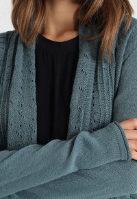 Kvinna iklädd en teal stickad kofta över en svart topp, med armarna korsade, synlig axellång ljusbrun frisyr.