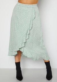 Vrouw in een groene bloemen midi wikkelrok met ruches, gecombineerd met zwarte soklaarzen, staand tegen een effen witte achtergrond.