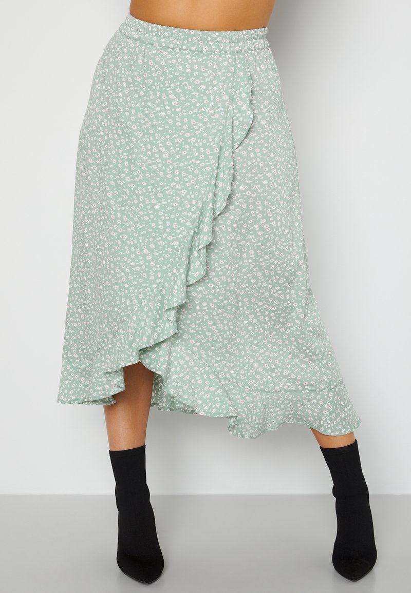 Vrouw in een groene bloemen midi wikkelrok met ruches, gecombineerd met zwarte soklaarzen, staand tegen een effen witte achtergrond.