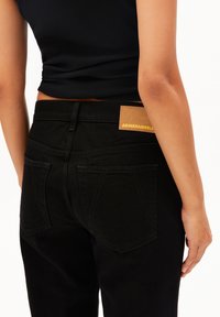 Pantalon en denim noir à taille haute, avec des poches arrières visibles et une étiquette en cuir beige portant "ARMEDANGELS" cousue à l'arrière. Texture lisse.