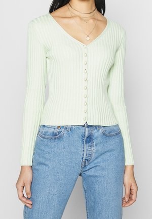 Vrouw die een lichtgroene geribbelde cardigan draagt met parelknopen, gecombineerd met lichtblauwe high-waisted jeans en gelaagde gouden kettingen.