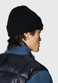 Czarna prążkowana czapka typu beanie wykonana z dzianiny, z podwiniętym rondem. Noszona z niebieską kurtką z polaru i czarną pikowaną kamizelką.