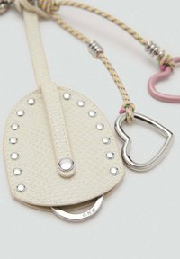 Petite pochette en cuir texturé crème avec des clous en métal, un anneau argenté et une corde tressée rose tenant un pendentif en forme de cœur en métal.
