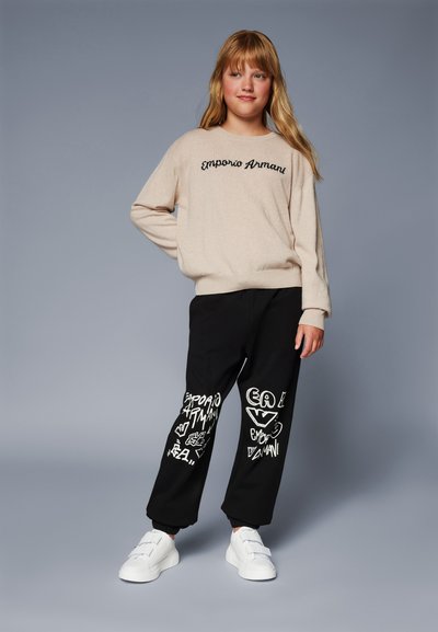 Beige kötött pulóver "Emporio Armani" logóval, fekete melegítőnadrággal kombinálva, amely fehér grafikai mintákkal és rugalmas mandzsettákkal rendelkezik. Fehér sportcipők.