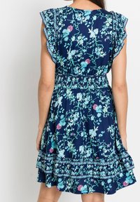 Vestido azul con estampado floral que presenta mangas con volantes, cintura fruncida y un dobladillo en capas con adornos decorativos. El material tiene una textura suave.