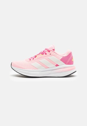 Chaussure de course rose clair avec des rayures blanches, des lacets roses, une semelle blanche rembourrée et une semelle extérieure noire sur un fond clair uni.