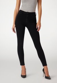 Jeans skinny negros de cintura alta con una textura suave, dos bolsillos frontales y cierre de botón. Combinados con tacones negros de punta afilada.