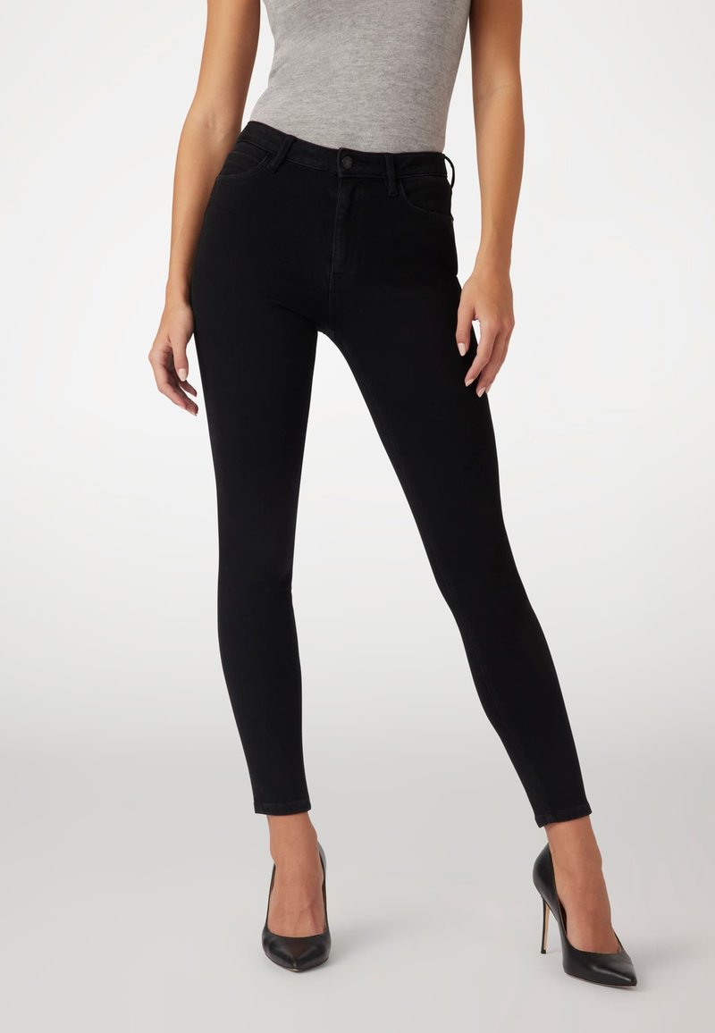 Jeans skinny negros de cintura alta con una textura suave, dos bolsillos frontales y cierre de botón. Combinados con tacones negros de punta afilada.