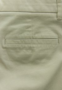 Khakifarbene Stofftasche auf der Rückseite der Hose, mit einem rechteckigen Paspeldesign und subtiler Textur sowie genähten Details entlang der Kanten.