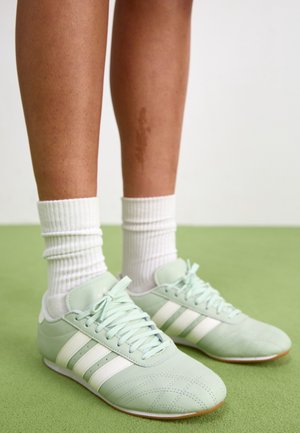 Jambes portant des chaussettes blanches côtelées aux chevilles et des baskets Adidas vert clair avec des bandes blanches, debout sur un sol vert.