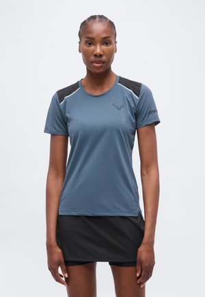 Γυναίκα που στέκεται μπροστά φοράει μπλε αθλητικό κοντομάνικο μπλουζάκι με μαύρες λεπτομέρειες και μαύρη αθλητική φούστα σε λευκό φόντο.