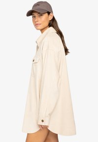 Chemise en velours côtelé beige avec un col, une poche avant, des poignets boutonnés et une coupe ample, associée à une casquette marron.