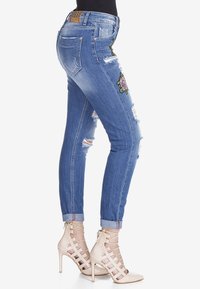 Jeans de denim azul ajustados con detalles desgastados y acentos florales bordados. Presenta un clásico diseño de cinco bolsillos y bajos marcados.