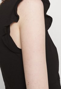 Haut noir avec manches à volants, tissu doux et coupe ajustée. Peau visible avec de légères taches de rousseur au niveau des épaules.