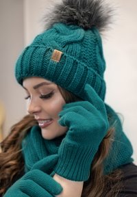Un bonnet tricot turquoise avec un pompon en fausse fourrure, des gants turquoise assortis et une écharpe texturée, avec un détail d'étiquette en bois sur le bonnet.
