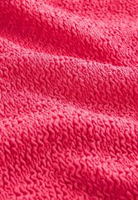 Lähikuva teksturoidusta vaaleanpunaisesta kankaasta, jossa on koholla olevia, aaltoilevia kuvioita ja pehmeä, tikattu ilme.