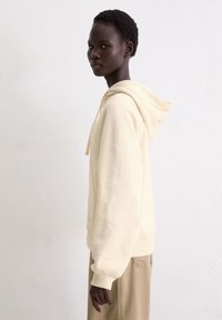 Crèmekleurige hoodie van een zacht materiaal met een trekkoord in de capuchon, ribbelboorden en onderrand, gedragen met beige broeken, zijwaarts bekeken.