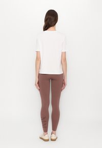 Hvit kortermet T-skjorte og brune leggings med grafisk aksent på venstre ben; modellen har på seg lette sko og har langt hår.