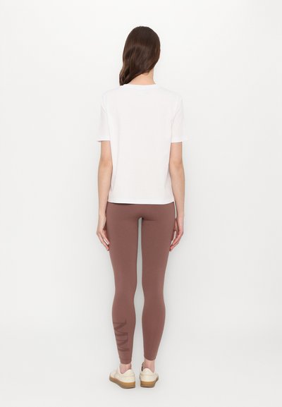 Bela kratka majica s kratkimi rokavi in rjave pajkice z grafičnim poudarkom na levi nogi; model nosi svetlo obutev in ima dolge lase.