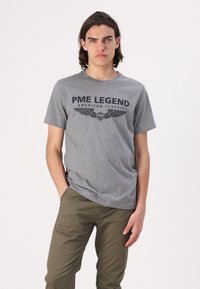 Šedé bavlněné tričko se silným černým potiskem "PME LEGEND AMERICAN CLASSIC" s křídly. Model má na sobě olivově zelené kalhoty.