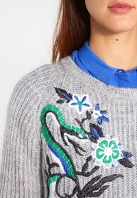 Pull gris en tricot orné d'une broderie florale et de serpent colorée, avec une chemise bleue vive à col visible en dessous. Détail en fil texturé apparent.