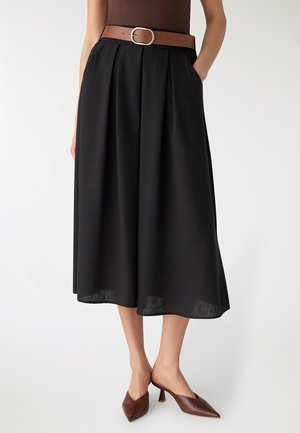 Donna che indossa una gonna midi nera a pieghe, una cintura marrone con fibbia dorata e mules con tacco marroni con punta appuntita, in piedi davanti a uno sfondo bianco.