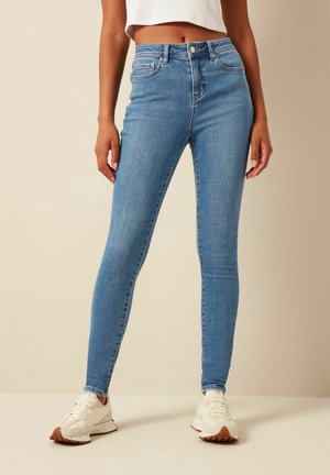Femme portant un jean skinny bleu clair et des baskets blanches, debout devant un fond beige.
