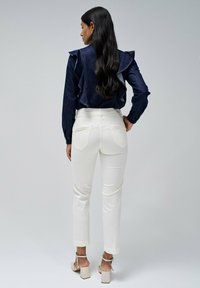 Top de denim con acentos de volantes, combinado con pantalones ajustados blancos. La modelo lleva sandalias de tacón cuadrado color beige. Diseño simple y limpio.