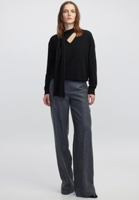 Zwarte longsleeve trui met een V-hals en een sjaaldetail, gecombineerd met grijze wijde broek, geplaatst tegen een effen achtergrond.