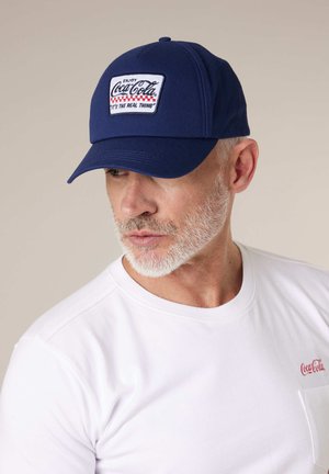 Homme à la barbe grise portant une casquette bleu marine avec le logo Coca-Cola et un t-shirt blanc avec un petit logo Coca-Cola sur la poitrine, regardant vers la gauche.