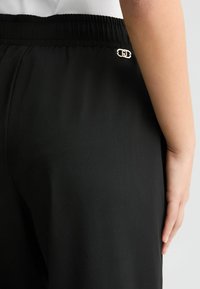 Zwarte broek met een elastische tailleband, met een metallic logoaccent. De stof is glad met een matte afwerking.