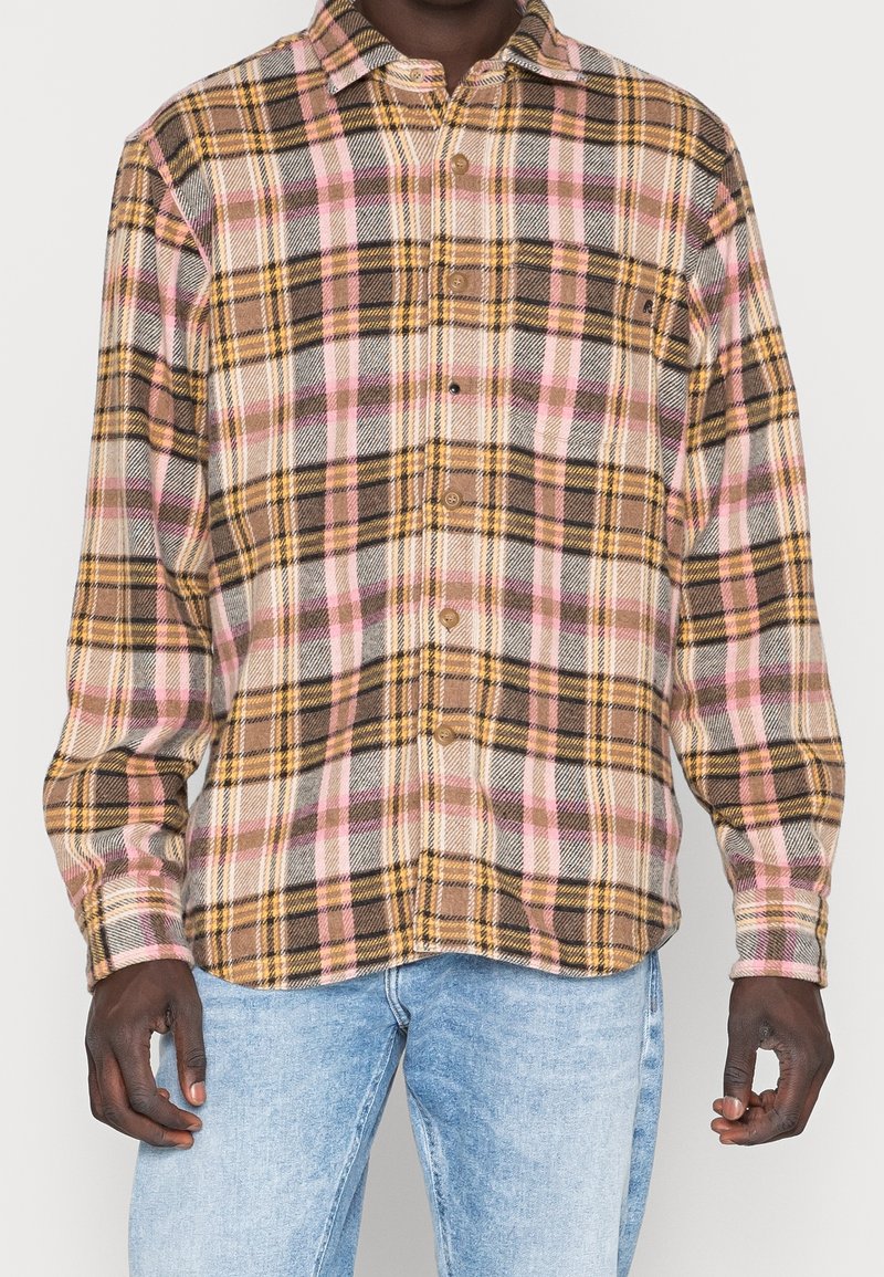 Homme portant une chemise à manches longues à boutons avec un motif à carreaux beige, marron, jaune et rose, associée à un jean bleu clair.