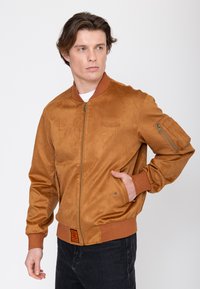 Mann trägt braune Wildleder-Bomberjacke mit Reißverschlusstasche am Ärmel und gerippten Bündchen, schaut seitlich und hat eine Hand in der Jackentasche.