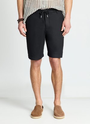 Mann trägt schwarze Shorts mit Kordelzug, braune Slipper, ein gemustertes Hemd und Armbänder und steht vor einem einfachen Hintergrund.