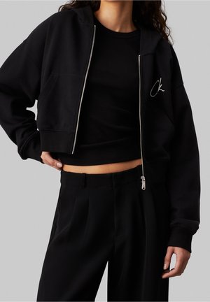 Kobieta ubrana w czarną krótko prążkowaną bluzę z zamkiem na tle jednolitego szarego tła, pod którą ma czarny prążkowany krótki top, oraz wysokie czarne spodnie.