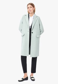 Cappotto di lana verde chiaro con una silhouette rilassata, rever a punta, chiusura con un unico bottone e tasche laterali. Abbinato a pantaloni neri e scarpe.