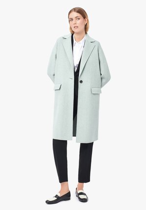 Lichtgroene wollen jas met een ontspannen silhouet, reverskraag, een enkele knopenclosure en zijzakken. Gepaard met zwarte broek en schoenen.