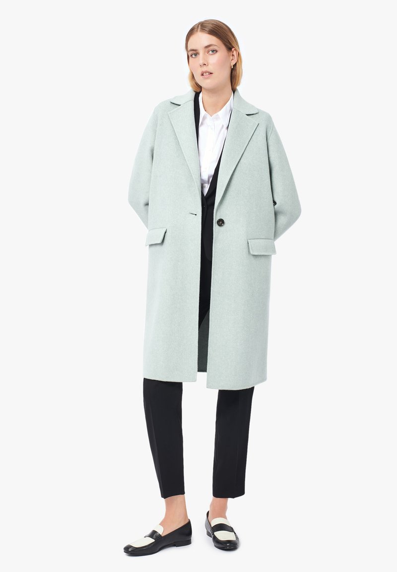 Cappotto di lana verde chiaro con una silhouette rilassata, rever a punta, chiusura con un unico bottone e tasche laterali. Abbinato a pantaloni neri e scarpe.