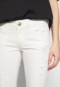 Jeans de denim blanco con un clásico diseño de 5 bolsillos, detalles desgastados en la parte frontal y un botón de metal dorado en la cintura.