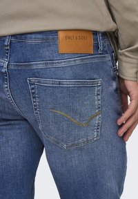 Μπλε τζιν με δερμάτινο ταμπελάκι με την επιγραφή "ONLY & SONS." Η πίσω τσέπη διαθέτει λεπτομέρεια με κίτρινη ραφή σε κυματιστό μοτίβο.