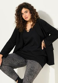 Zwarte blazer over een zwarte top, gecombineerd met grijze leggings met dierenprintpatronen. De leggings hebben verschillende texturen en ontwerpen.