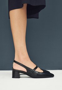 Tacones slingback de piel negra con puntera afilada, tacón bajo y bloque, y una tira delgada que cruza el pie; textura suave y diseño minimalista.