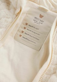 Innenfutter des Babybekleidungsstücks aus Fleece in Creme mit Pflegeetikett. Das Etikett zeigt Bärengrafiken und Platz für vier Namen. Mit Reißverschluss.