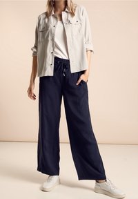 Kvinna som bär en ljusbeige knapp-up-skjorta över en vit topp, marinblå vida byxor med dragstring och vita sneakers, stående mot en beige bakgrund.