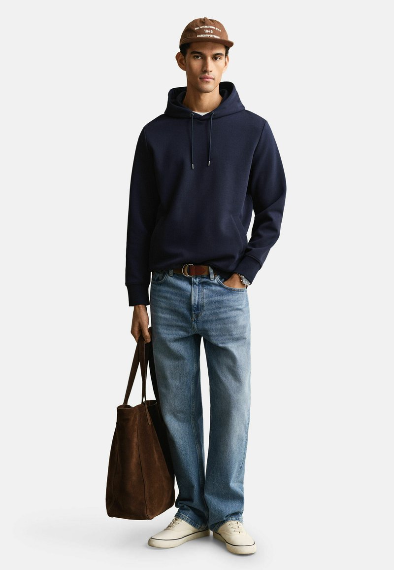 Junger Mann trägt eine marineblaue Kapuzenjacke, hellblaue Jeans, weiße Turnschuhe, braune Mütze und trägt eine große braune Wildledertasche.