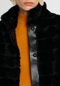 Manteau en fausse fourrure noire au toucher doux, avec un devant en cuir noir brillant et des boutons ronds noirs, porté sur un pull orange à côtes.