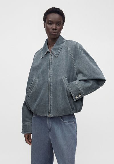 Personne portant une veste en jean délavée oversize avec un large col et un pantalon en jean assorti sur un fond clair uni.