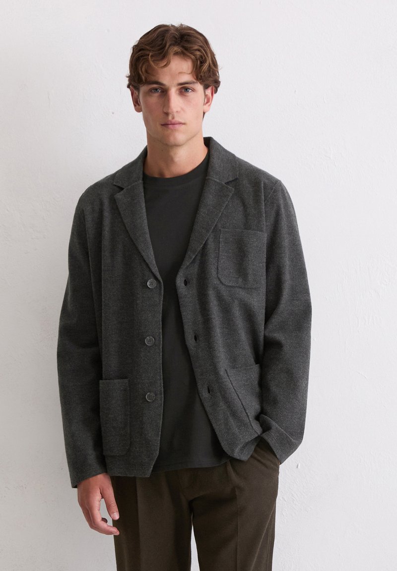 Grijze wollen blazer met een reverskraag, twee voorzakken en knoopsluiting, gedragen over een zwart overhemd, met een ontspannen pasvorm.