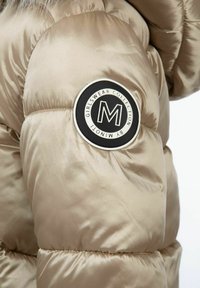 Beige quilted jakke med en glansfuld overflade. Har et rundt sort mærke på ærmet med hvid skrift og et fremtrædende "M" logo.