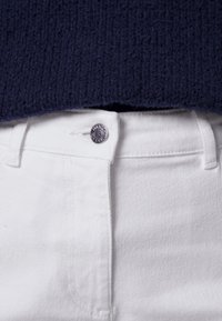 Close-up van de taille van witte spijkerbroek met zilverkleurige knoop en riemlussen, gecombineerd met de zoom van een donkerblauwe gebreide trui.